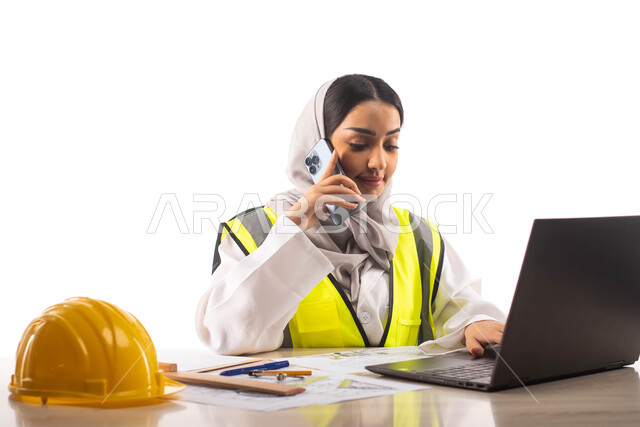 اجراء مكالمة صوتية عبر الهاتف المحمول ، ارتداء بدلة السلامة, بورتريه لمهندسة معمارية عربية خليجية سعودية محترفه داخل مكتب هندسي ، إستخدام جهاز الحاسوب المحمول للإنجاز وتتبع المهام ، خوذة الوقاية الصفراء ،مهن ووظائف ، خلفية بيضاء