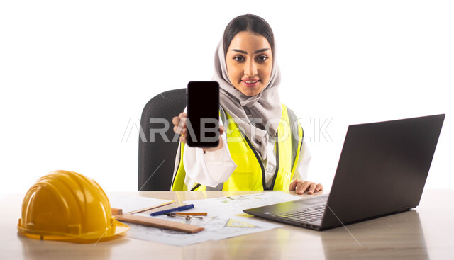 ارتداء سترة الحماية الخاصة بالعمل، عرض شاشة سوداء فارغة عبر الهاتف المحمول, ادارة المشاريع الهندسية، بورتريه لمهندسة معمارية عربية خليجية سعودية مبتسمة تجلس في المكتب , استخدام الحاسوب المحمول , مخططات و تصماميم هندسية