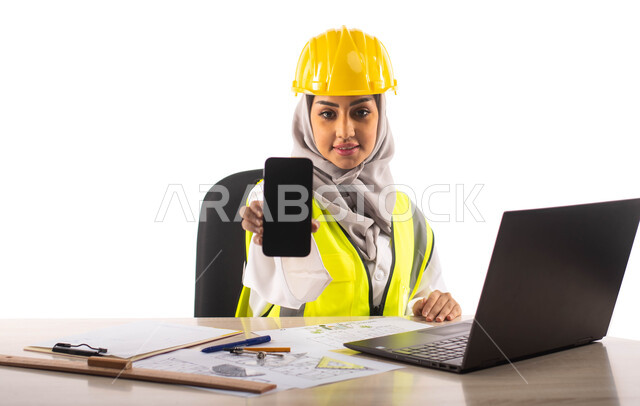 عرض شاشة سوداء فارغة عبر الهاتف المحمول, ادارة المشاريع الهندسية، بورتريه لمهندسة معمارية عربية خليجية سعودية مبتسمة تجلس في المكتب ,ارتداء خوذة وسترة الحماية الخاصة بالعمل، استخدام الحاسوب المحمول , مخططات و تصماميم هندسية