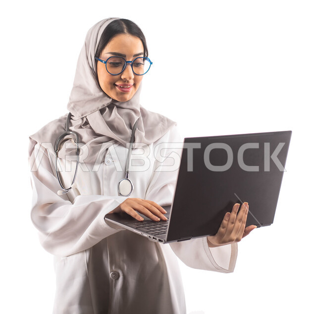  مفهوم إستخدام أجهزة تقنية حديثة, ايماءات تدل على السعادة,  العمل في القطاع الصحي, بورتريه لطبيبة عربية سعودية خليجية محجبة  تحمل بيدها الحاسوب المحمول , تقديم الخدمات للمرضى بكل كفاءة ،ارتداء المعطف الطبي والنظارات الطبية, مفهوم الطب والرعاية الصحية ، خل