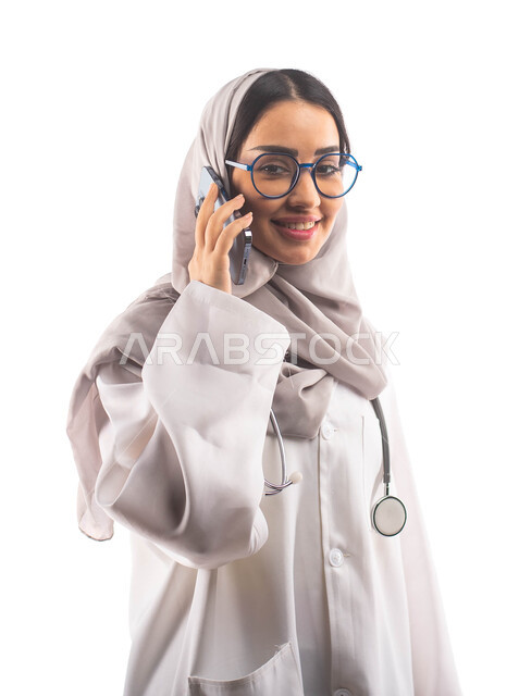 ايماءات تدل على السعادة, اجراء مكالمة صوتية عبر الجوال,  مفهوم إستخدام أجهزة تقنية حديثة, العمل في القطاع الصحي, بورتريه لطبيبة عربية سعودية خليجية محجبة ومبتسمة, تقديم الخدمات للمرضى بكل كفاءة ،ارتداء المعطف الطبي والنظارات الطبية, مفهوم الطب والرعاية ال