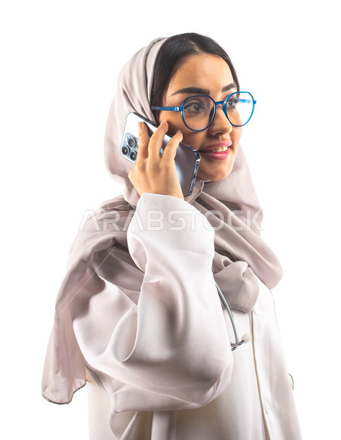 اجراء مكالمة صوتية عبر الجوال,  مفهوم إستخدام أجهزة تقنية حديثة, العمل في القطاع الصحي, بورتريه لطبيبة عربية سعودية خليجية محجبة ومبتسمة, تقديم الخدمات للمرضى بكل كفاءة ،ارتداء المعطف الطبي والنظارات الطبية, مفهوم الطب والرعاية الصحية ، مهن و وظائف ، خلفي