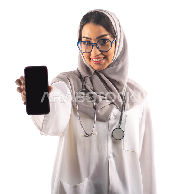 عرض شاشة سوداء فارغة عبر الجوال, العمل في القطاع الصحي, بورتريه لطبيبة عربية سعودية خليجية محجبة ومبتسمة, تقديم الخدمات للمرضى بكل كفاءة ،ارتداء المعطف الطبي والنظارات الطبية, مفهوم الطب والرعاية الصحية ، مهن و وظائف ، خلفية بيضاء