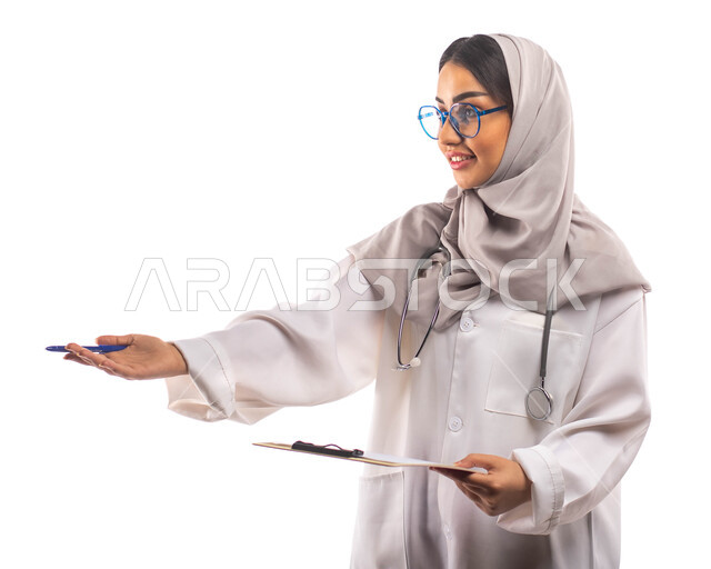 العمل في القطاع الصحي, ايماءات تدل على الاستقبال, بورتريه لطبيبة عربية سعودية خليجية محجبة ومبتسمة, تحمل بيدها ملف خاص بالمرضى، ارتداء المعطف الطبي والنظارات الطبية, مفهوم الطب والرعاية الصحية ، مهن و وظائف  ، خلفية بيضاء