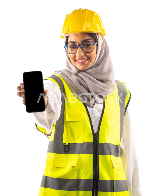 مهن ووظائف ،عرض شاشة سوداء فارغة عبر الهاتف المحمول, ايماءات تدل على السعادة والفرح, بورتريه لمهندسة انشاءات معمارية عربية سعودية خليجية مبتسمة ترتدي النظارات الطبية ، ارتداءالخوذة وبدلة السلامة الخاصة في العمل ،مخططات وتصاميم هندسية،  خلفية بيضاء