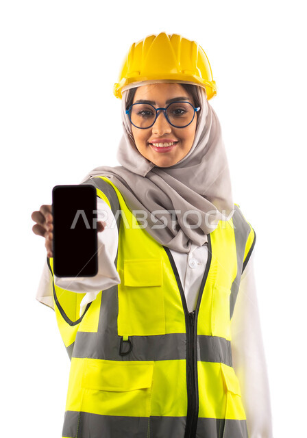 عرض شاشة سوداء فارغة عبر الهاتف المحمول, ايماءات تدل على السعادة والفرح, بورتريه لمهندسة انشاءات معمارية عربية سعودية خليجية مبتسمة ترتدي النظارات الطبية ، ارتداءالخوذة وبدلة السلامة الخاصة في العمل ،مخططات وتصاميم هندسية، مهن ووظائف ،  خلفية بيضاء