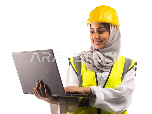 تحمل بيدها الحاسوب المحمول, ايماءات تدل على السرور والفرح, بورتريه  لمهندسة انشاءات معمارية عربية سعودية خليجية مبتسمة،  ارتداءالخوذة وبدلة السلامة الخاصة في العمل ،مخططات وتصاميم هندسية، مهن ووظائف ، خلفية بيضاء