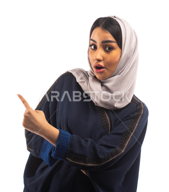 تعابير وجه تدل على الاندهاش والمفاجأة، بورتريه لامرأة عربية خليجية سعودية محجبة ترتدي العباءة السوداء و تشير باصبع السبابة نحو شيء ما، حركة اصبع واشارات، خلفية بيضاء