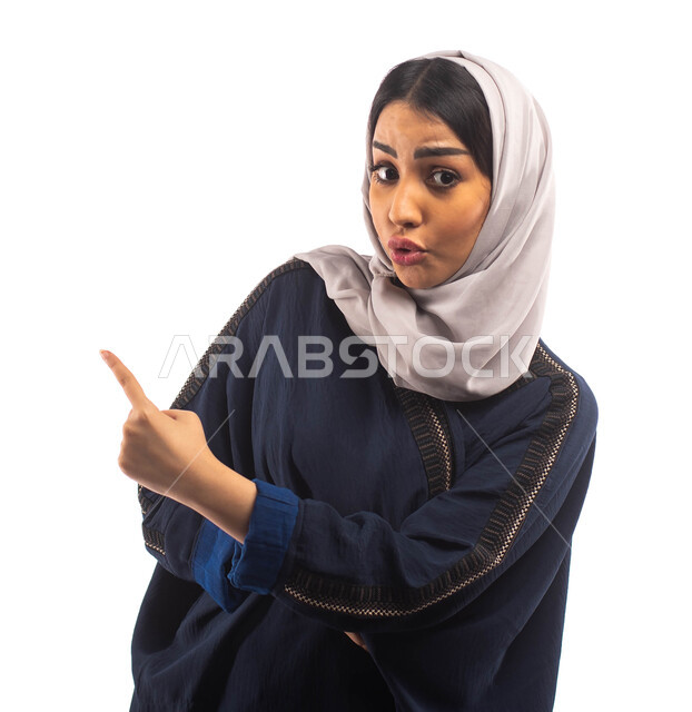 تعابير وجه تدل على المفاجأة و الاندهاش، بورتريه لامرأة عربية خليجية سعودية محجبة ترتدي العباءة السوداء و تشير باصبع السبابة نحو شيء ما، حركة اصبع واشارات، خلفية بيضاء