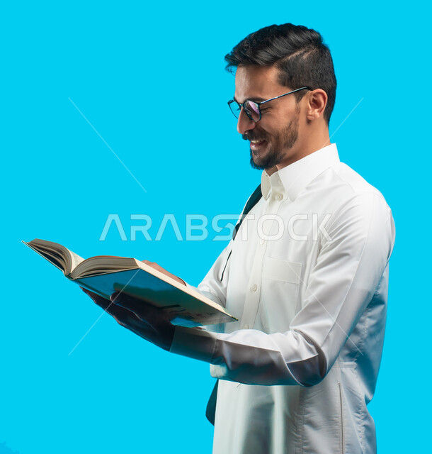 التعليم الجامعي في المملكة العربية السعودية ، إيماءات وجه ويدين تدل على الفرح والسعادة ، تدوين الملاحظات وكتابة تقارير وأبحاث علمية ، بورتريه لطالب جامعي عربي سعودي يرتدي الزي الخليجي و يحمل بيده كتب ، مراجعة الدروس اليومية ، خلفية زرقاء
