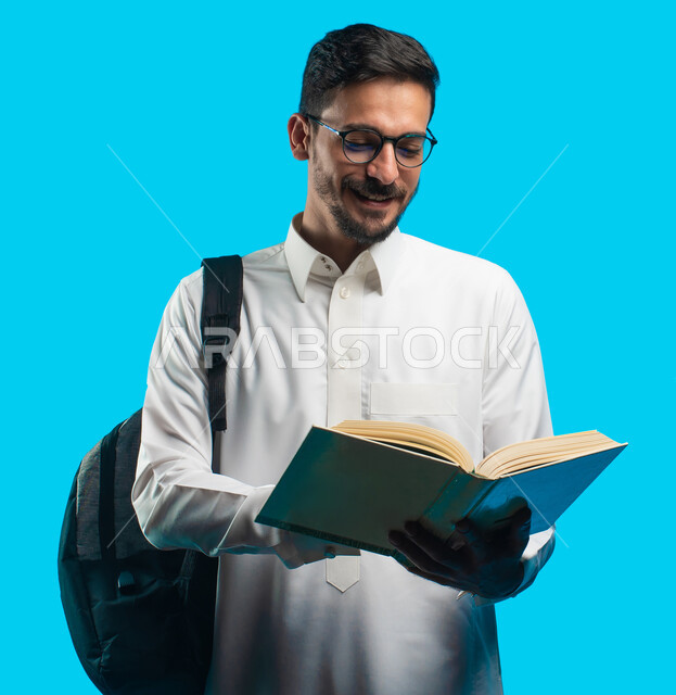  تدوين الملاحظات وكتابة تقارير وأبحاث علمية ، التعليم الجامعي في المملكة العربية السعودية ، إيماءات وجه ويدين تدل على الفرح والإعجاب ،بورتريه لطالب جامعي عربي سعودي يرتدي الزي الخليجي و يحمل بيده كتب ، مراجعة الدروس اليومية ، خلفية زرقاء