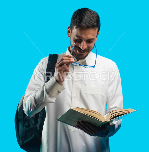 مراجعة الدروس اليومية ، التعليم الجامعي في المملكة العربية السعودية ، إيماءات وجه ويدين تدل على الفرح والإعجاب ، تدوين الملاحظات وكتابة تقارير وأبحاث علمية ، بورتريه لطالب جامعي عربي سعودي يرتدي الزي الخليجي و يحمل بيده كتب ، خلفية زرقاء