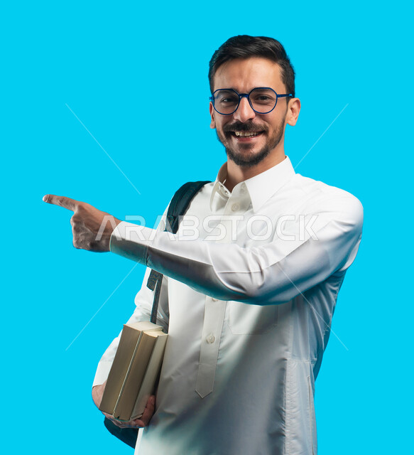 تدوين الملاحظات وكتابة تقارير وأبحاث علمية ، التعليم الجامعي في المملكة العربية السعودية ، إيماءات وجه ويدين تدل على الفرح والإعجاب ، بورتريه طالب جامعي عربي سعودي يرتدي الزي الخليجي و يشير بإصبعه لبعيد ، مراجعة الدروس اليومية ، خلفية زرقاء