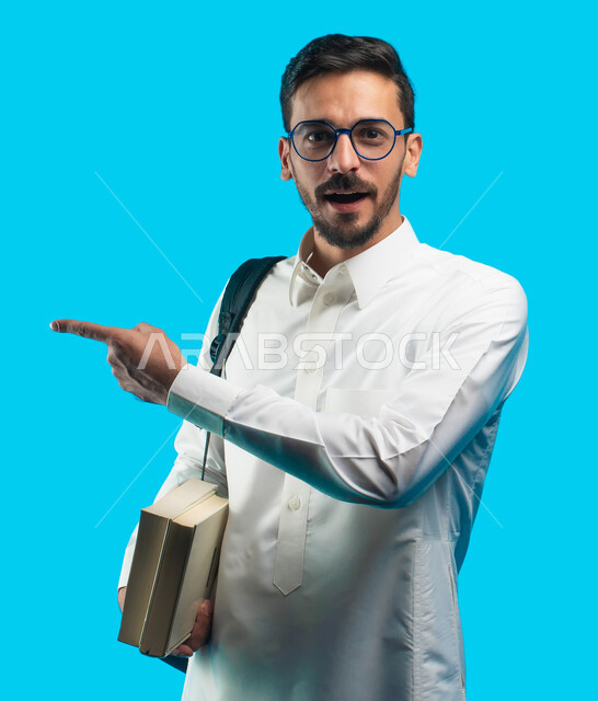مراجعة الدروس اليومية ، التعليم الجامعي في المملكة العربية السعودية ، إيماءات وجه ويدين تدل على الدهشة ، تدوين الملاحظات وكتابة تقارير وأبحاث علمية ، بورتريه طالب جامعي عربي سعودي يرتدي الزي الخليجي و يشير يإصبعه لبعيد ، خلفية زرقاء
