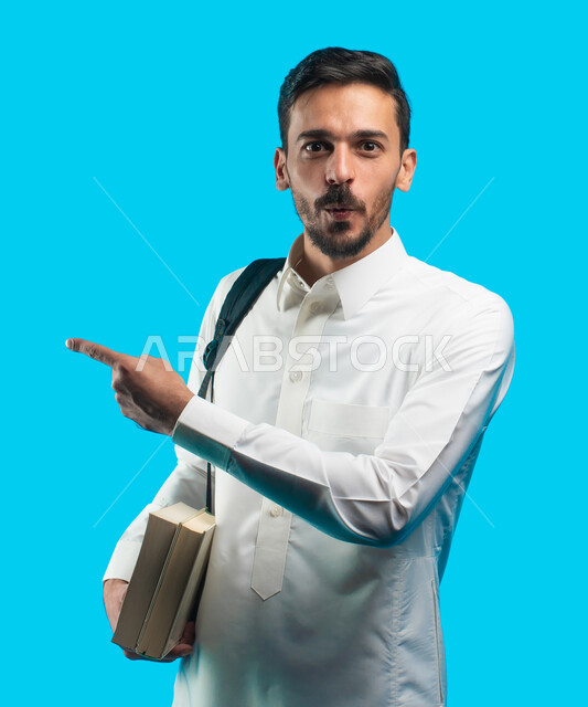 التعليم الجامعي في المملكة العربية السعودية ، إيماءات وجه ويدين تدل على الدهشة ، تدوين الملاحظات وكتابة تقارير وأبحاث علمية ، بورتريه طالب جامعي عربي سعودي يرتدي الزي الخليجي و يحمل بيده كتب ويشير بإصبعه لبعيد ، مراجعة الدروس اليومية ، خلفية زرقاء