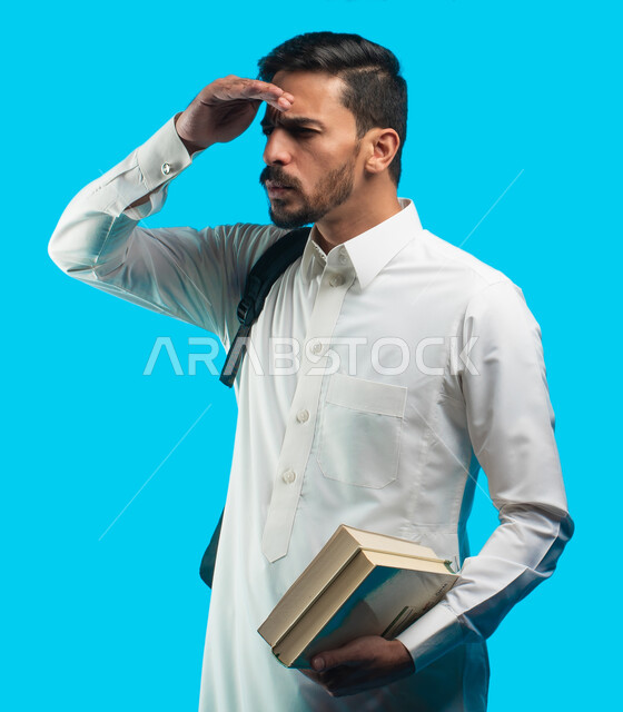 التعليم الجامعي في المملكة العربية السعودية ، إيماءات وجه ويدين ينظر لبعيد بإستغراب ، كتابة تقارير وأبحاث علمية ، بورتريه طالب جامعي عربي سعودي يرتدي الزي الخليجي و يحمل بيده كتب ، مراجعة الدروس اليومية ، خلفية زرقاء