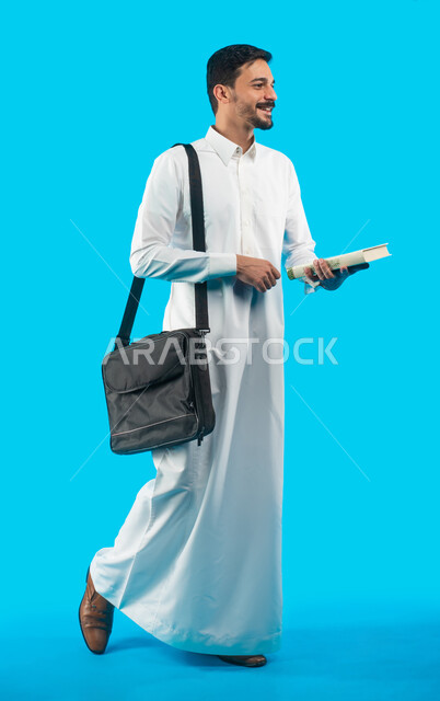 Education in the Kingdom of Saudi Arabia, reviewing daily lessons, full-length body image, facial gestures indicating joy and success, portrait of a university student wearing the Saudi Gulf uniform holding books in his hand, taking notes and writing scie