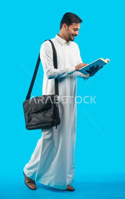 مراجعة الدروس اليومية ، صورة الجسم بالطول الكامل ، إيماءات وجه تدل على الفرح والنجاح ، بورتريه طالب جامعي يرتدي الزي الخليجي السعودي يحمل بيده كتب ، تدوين الملاحظات و كتابة تقارير وأبحاث علمية ، التعليم في المملكة العربية السعودية ، خلفية زرقاء