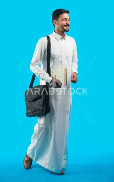 صورة الجسم بالطول الكامل ، مراجعة الدروس اليومية ، إيماءات وجه تدل على الفرح والنجاح ، بورتريه طالب جامعي يرتدي الزي الخليجي السعودي يحمل بيده كتب ، تدوين الملاحظات و كتابة تقارير وأبحاث علمية ، التعليم في المملكة العربية السعودية ، خلفية زرقاء