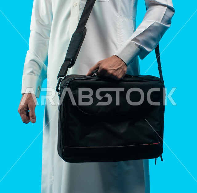 الإستعداد للذهاب إلى العمل , بورتريه لرجل أعمال عربي خليجي سعودي يرتدي الزي السعودي يحمل حقيبة كمبيوتر محمول على كتفه طالب جامعي عربي سعودي يستعد للذهاب للجامعة ، أداء الواجبات والدروس عبر الحاسوب , خلفية زرقاء