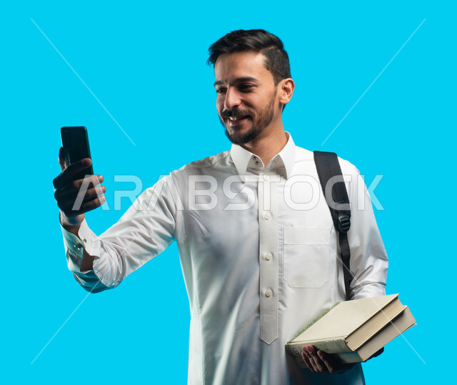التعليم الجامعي في المملكة العربية السعودية ، إستخدام أجهزة تقنية حديثة ، إيماءات وجه تدل على الفرح بنتيجة الاختبارات النهائية ، بورتريه لطالب جامعي عربي يرتدي الزي الخليجي السعودي يقوم بمكالمة فيديو من خلال الهاتف المحمول ، خلفية زرقاء