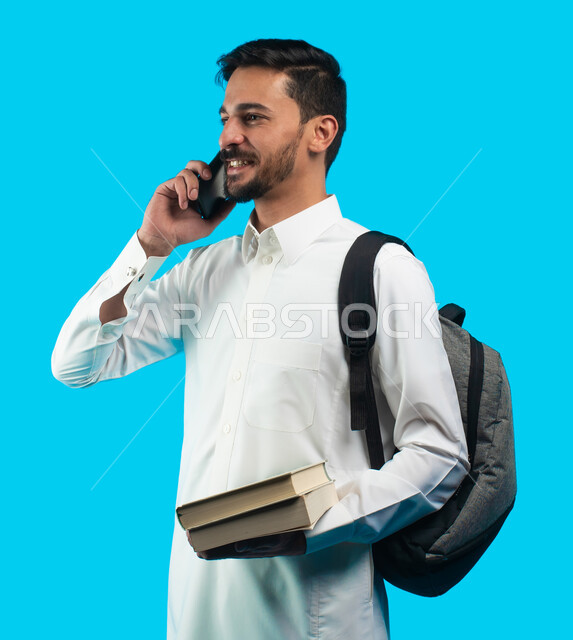 إيماءات وجه تدل على الفرح بنتيجة الاختبارات النهائية ، بورتريه لطالب جامعي عربي يرتدي الزي الخليجي السعودي يتحدث على الهاتف المحمول حول نتائج الإمتحان ، شهادة التفوق والنجاح والتقدم ، التعليم الجامعي في المملكة العربية السعودية ، خلفية زرقاء