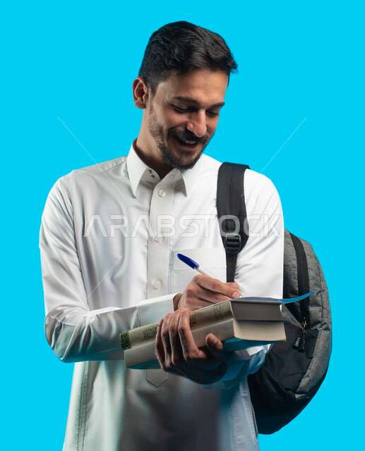 إيماءات تدل على السعادة بإجتياز الإمتحان ، بورتريه لطالب جامعي عربي يرتدي الزي الخليجي السعودي و يحمل بيده أوراق الإختبار ، مراجعة الدروس اليومية ، التعليم الجامعي في المملكة العربية السعودية ، تدوين الملاحظات وكتابة تقارير أبحاث علمية ، خلفية زرقاء