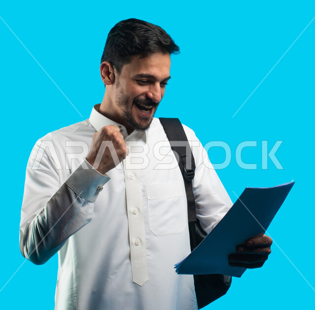  تدوين الملاحظات وكتابة تقارير أبحاث علمية ، إيماءات تدل على السعادة بإجتياز الإمتحان ، بورتريه لطالب جامعي عربي يرتدي الزي الخليجي السعودي و يحمل بيده أوراق الإختبار ، مراجعة الدروس اليومية ، التعليم الجامعي في المملكة العربية السعودية ، خلفية زرقاء