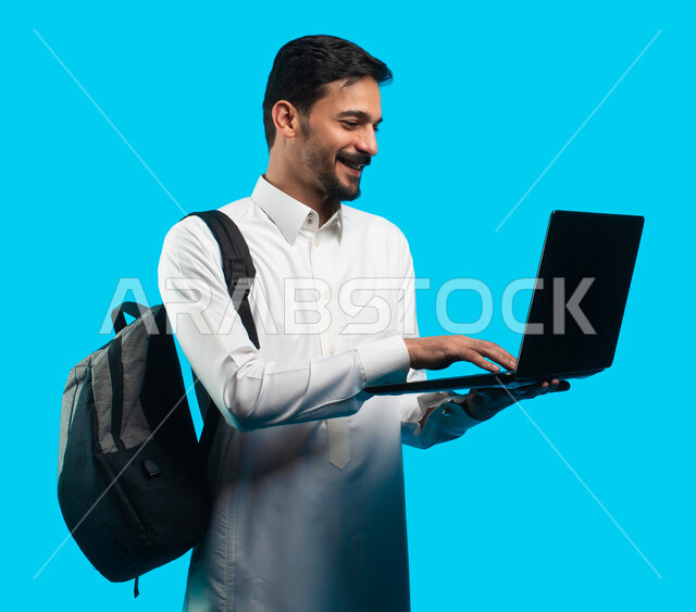 إيماءات تدل على السعادة ، إستخدام أجهزه تقنية حديثة ،  بورتريه لطالب جامعي عربي يرتدي الزي الخليجي السعودي ويقوم بإجراء مكالمة فيديو من خلال كمبيوتر محمول ، الإستعداد للمحاضرات الجامعية الالكترونية ، التعلم عن بعد ، مراجعة الدروس اليومية ، خلفية زرقاء