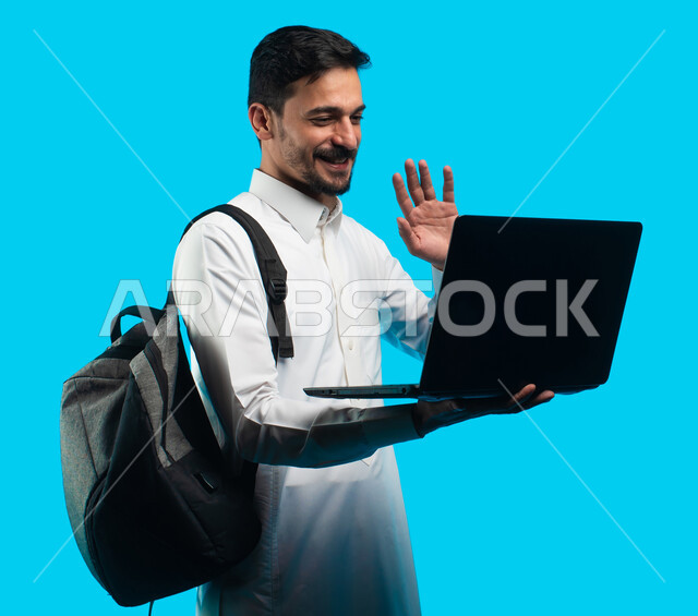 إستخدام أجهزه تقنية حديثة ، إيماءات تدل على السعادة ، بورتريه لطالب جامعي عربي يرتدي الزي الخليجي السعودي ويقوم بإجراء مكالمة فيديو من خلال كمبيوتر محمول ، الإستعداد للمحاضرات الجامعية الالكترونية ، التعلم عن بعد ، مراجعة الدروس اليومية ، خلفية زرقاء