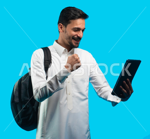 Portrait of an Arab university student wearing the Saudi Gulf uniform making a video call through a tablet device, facial gestures indicating happiness, preparation for electronic lectures, distance learning, reviewing daily lessons, education in the King