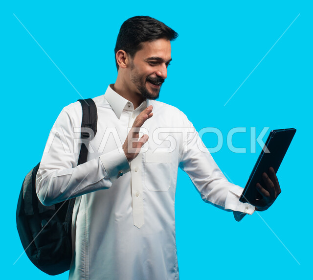 إيماءات وجه تدل على السعادة ، بورتريه لطالب جامعي عربي يرتدي الزي الخليجي السعودي يقوم بإجراء مكالمة فيديو من خلال جهاز التابلت ، الإستعداد للمحاضرات الالكترونية ، التعلم عن بعد ، مراجعة الدروس اليومية ، التعليم في المملكة العربية السعودية ، خلفية زرقاء