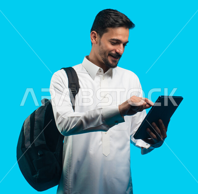  إستخدام أجهزة تقنية حديثة ، بورتريه لطالب جامعي عربي يرتدي الزي الخليجي السعودي يقوم بإجراء مكالمة فيديو من خلال جهاز التابلت ، الإستعداد للمحاضرات الالكترونية ، التعلم عن بعد ، مراجعة الدروس اليومية ، التعليم في المملكة العربية السعودية ، خلفية زرقاء