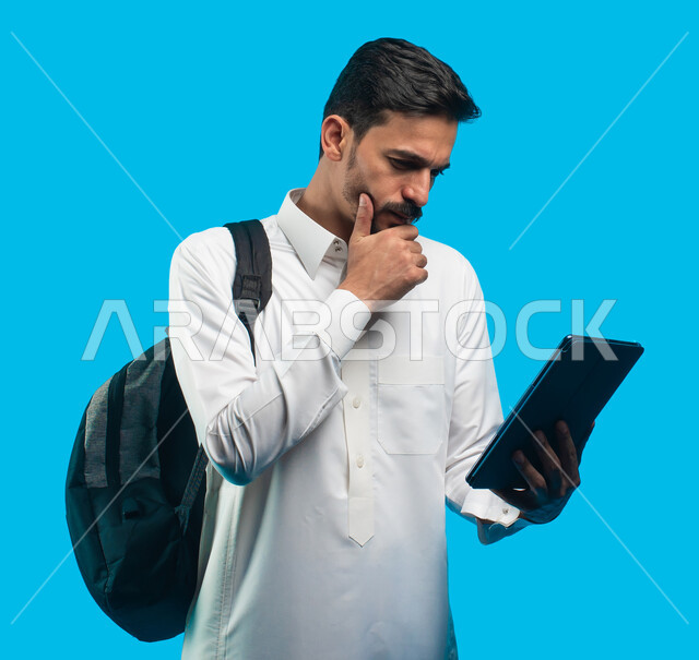 التعلم عن بعد ، إيماءات وجه ويدين تدل على التفكير والحيرة ، بورتريه لطالب جامعي عربي يرتدي الزي الخليجي السعودي يقوم بإجراء مكالمة فيديو من خلال جهاز التابلت ، الإستعداد للمحاضرات الجامعية الالكترونية ، التعليم في المملكة العربية السعودية ، خلفية زرقاء