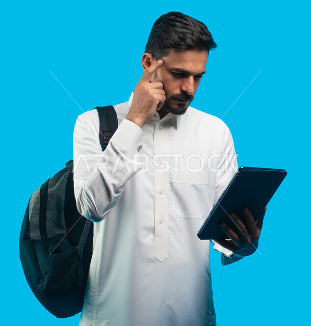 التعليم في المملكة العربية السعودية ، إيماءات وجه ويدين تدل على التفكير والحيرة ، بورتريه لطالب جامعي عربي يرتدي الزي الخليجي السعودي يقوم بإجراء مكالمة فيديو من خلال جهاز التابلت ، الإستعداد للمحاضرات الجامعية الالكترونية ، التعلم عن بعد ، خلفية زرقاء