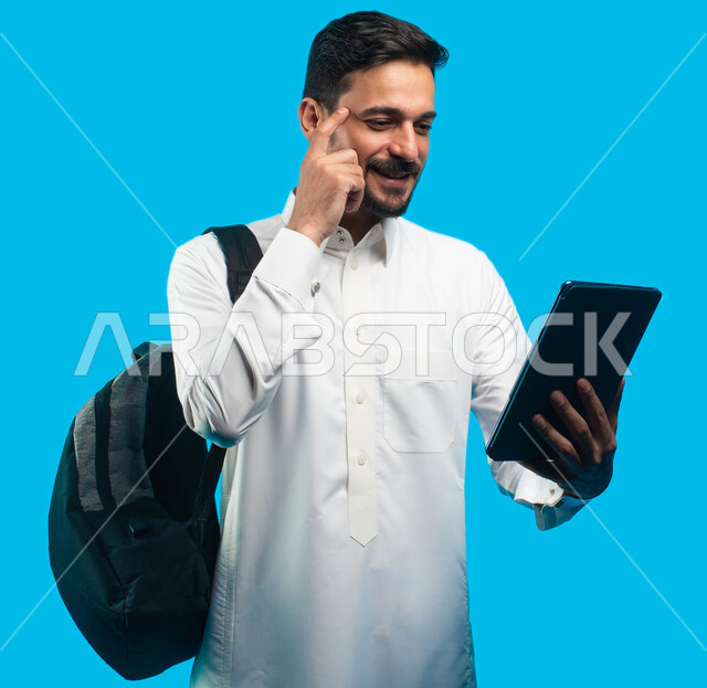 إيماءات وجه ويدين تدل على التفكير ، بورتريه لطالب جامعي عربي يرتدي الزي الخليجي السعودي يقوم بإجراء مكالمة فيديو من خلال جهاز التابلت ، الإستعداد للمحاضرات الجامعية الالكترونية ، التعلم عن بعد ، التعليم في المملكة العربية السعودية ، خلفية زرقاء