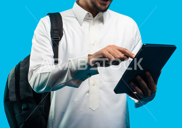 الإستعداد للمحاضرات الالكترونية ، إستخدام أجهزة تقنية حديثة ، بورتريه لطالب جامعي عربي يرتدي الزي الخليجي السعودي يقوم بإجراء مكالمة فيديو من خلال جهاز التابلت ، التعلم عن بعد ، مراجعة الدروس اليومية ، التعليم في المملكة العربية السعودية ، خلفية زرقاء
