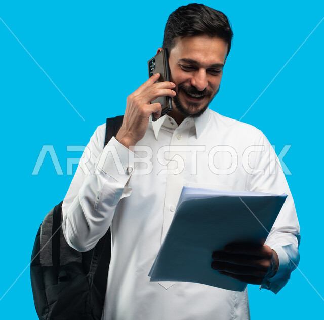 بورتريه لطالب جامعي عربي يرتدي الزي الخليجي السعودي يتحدث على الهاتف المحمول حول نتائج الإمتحان ، شهادة التفوق والنجاح والتقدم ، إيماءات وجه تدل على الفرح بنتيجة الاختبارات النهائية ، التعليم الجامعي في المملكة العربية السعودية ، خلفية زرقاء