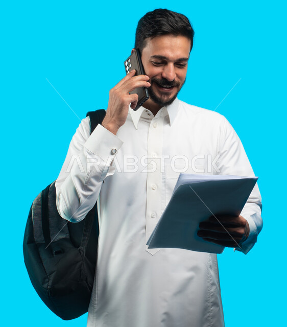 شهادة التفوق والنجاح والتقدم ، بورتريه لطالب جامعي عربي يرتدي الزي الخليجي السعودي يتحدث على الهاتف المحمول حول نتائج الإمتحان ، إيماءات وجه تدل على الفرح بنتيجة الاختبارات النهائية ، التعليم الجامعي في المملكة العربية السعودية ، خلفية زرقاء