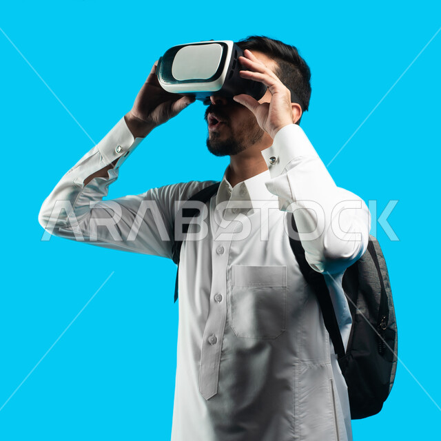 التفاعل و المتعة و التسلية بمشاهدة الأفلام ، مشاهدة أفلام ثلاثية الابعاد عبر نظارة الواقع الإفتراضي الحديث ، بورتريه شاب عربي خليجي سعودي يرتدي نظارات العالم الخيالي , التطور التقني والتكنولوجي , إيماءات وجه ويدين تدل على الحماس  , خلفيه زرقاء