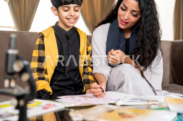 تلميذ سعودي خليجي يدرس في المنزل مع والدته ، تساعده على تأدية واجباته المدرسية ، التعلم عن بعد ، مفهوم التعليم عبر الانترنت ، التعليم المنزلي اثناء الحجر الصحي و تفشي فيروس كورونا ، العزلة الذاتية .