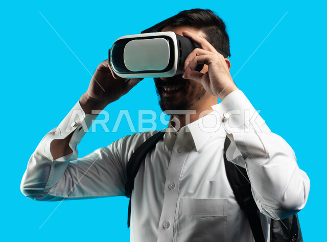 مشاهدة أفلام ثلاثية الابعاد عبر نظارة الواقع الإفتراضي الحديث ، التفاعل و المتعة و التسلية بمشاهدة الأفلام ، بورتريه شاب عربي خليجي سعودي يرتدي نظارات العالم الخيالي , التطور التقني والتكنولوجي , إيماءات وجه ويدين تدل على الحماس  , خلفيه زرقاء