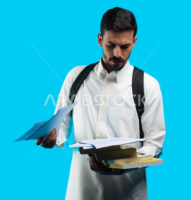 تدوين الملاحظات وكتابة تقارير وأبحاث علمية ، التعليم الجامعي في المملكة العربية السعودية ، إيماءات وجه ويدين تدل على التذمر والشعور بالإحباط والحزن من الضغط الدراسي ، بورتريه لطالب جامعي عربي يرتدي الزي الخليجي السعودي و يحمل بيده كتب ، خلقية زرقاء