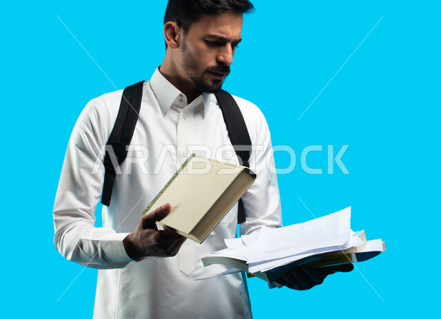 بورتريه لطالب جامعي عربي يرتدي الزي الخليجي السعودي و يحمل بيده كتب ، تدوين الملاحظات وكتابة تقارير وأبحاث علمية ، التعليم الجامعي في المملكة العربية السعودية ، إيماءات وجه ويدين تدل على التذمر والشعور بالإحباط والحزن من الضغط الدراسي ، خلفية زرقاء