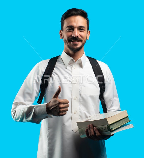 التعليم الجامعي في المملكة العربية السعودية ، إيماءات وجه ويدين تدل على الفرح والإعجاب ، تدوين الملاحظات وكتابة تقارير وأبحاث علمية ، بورتريه طالب جامعي عربي سعودي يرتدي الزي الخليجي و يحمل بيده كتب وعلى ظهره حقيبة ،مراجعة الدروس اليومية ، خلفية زرقاء