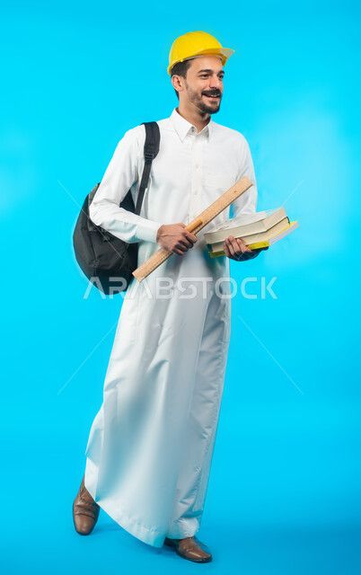 صورة الجسم بالطول الكامل ، إيماءات وجه تدل على الفرح ، بورتريه طالب جامعي يرتدي الزي الخليجي السعودي يحمل بيده كتب ومعدات الهندسة ، تدوين الملاحظات وكتابة تقارير وأبحاث علمية ، إدارة المشاريع الهندسية ، التعليم في المملكة العربية السعودية ، خلفية زرقاء 