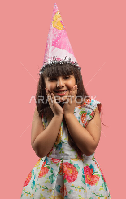 حركة يد وإشارات، بورتريه لفتاة عربية سعودية خليجية ترتدي قبعة الإحتفال بعيد الميلاد وتضع يديها على وجهها، قضاء أوقات ممتعة، إيماءات وجه ويدين تدل على الدهشة والتعجب، خلفية وردية