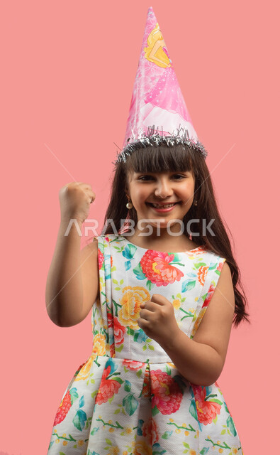 Hand movement and gestures indicating enthusiasm, holidays and happy occasions, a portrait of a Saudi Arabian Gulf girl wearing an elegant floral dress and birthday celebration hat, happy facial gestures, pink background
