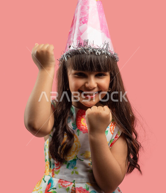 إيماءات وجه سعيدة، الأعياد والمناسبات السعيدة، بورتريه لفتاة عربية سعودية خليجية ترتدي فستان مورد أنيق وقبعة الإحتفال بعيد الميلاد، حركة يد وإشارات تدل على الحماس، خلفية وردية