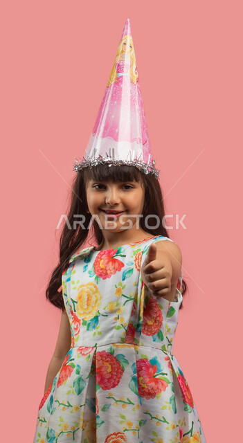 إيماءات وجه ويدين تدل على الإعجاب والقبول والموافقة، بورتريه لفتاة عربية خليجية سعودية ترتدي فستان مورد أنيق وقبعة الإحتفال بعيد الميلاد وترفع أصبع الإبهام لأعلى، أعياد ومناسبات سعيدة، حركة أصبع وإشارات، خلفية وردية